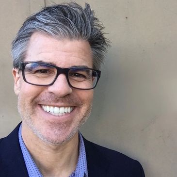 Smiling man with glasses and gray hair against a plain background.
