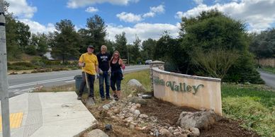 Three people standing by a renovated Green Valley sign near a roadside.