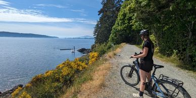 Woman with bicycle overlooking calm lake and yellow flowers on a sunny day.