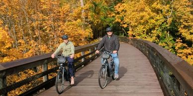 Two people biking on a wooden bridge surrounded by autumn foliage.