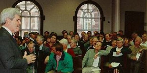 A speaker addressing an attentive audience in a room with large arched windows.