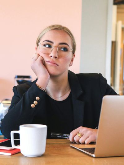 A woman looks thoughtfully away while sitting at a desk with a laptop and coffee.