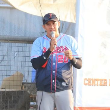 Man speaking into a microphone at an outdoor event, wearing a baseball jersey.
