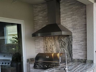 Outdoor kitchen that has a vent hood with natural stone siding on the wall. Same stone as kitchen