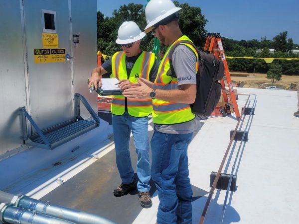 Two construction workers in safety gear review plans on a rooftop.