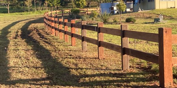 Curved wooden fence alongside a rural house and lush greenery.