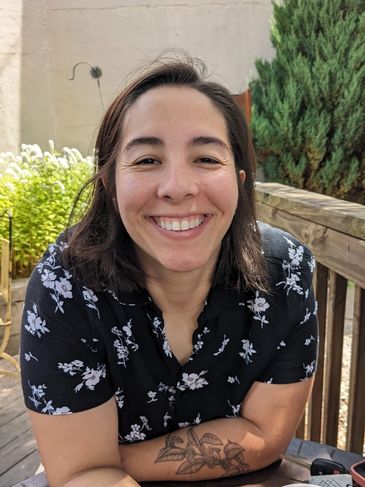 Smiling woman with floral shirt and a leaf tattoo on her forearm.