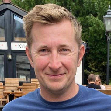 Smiling man outdoors with wooden chairs and greenery in background.