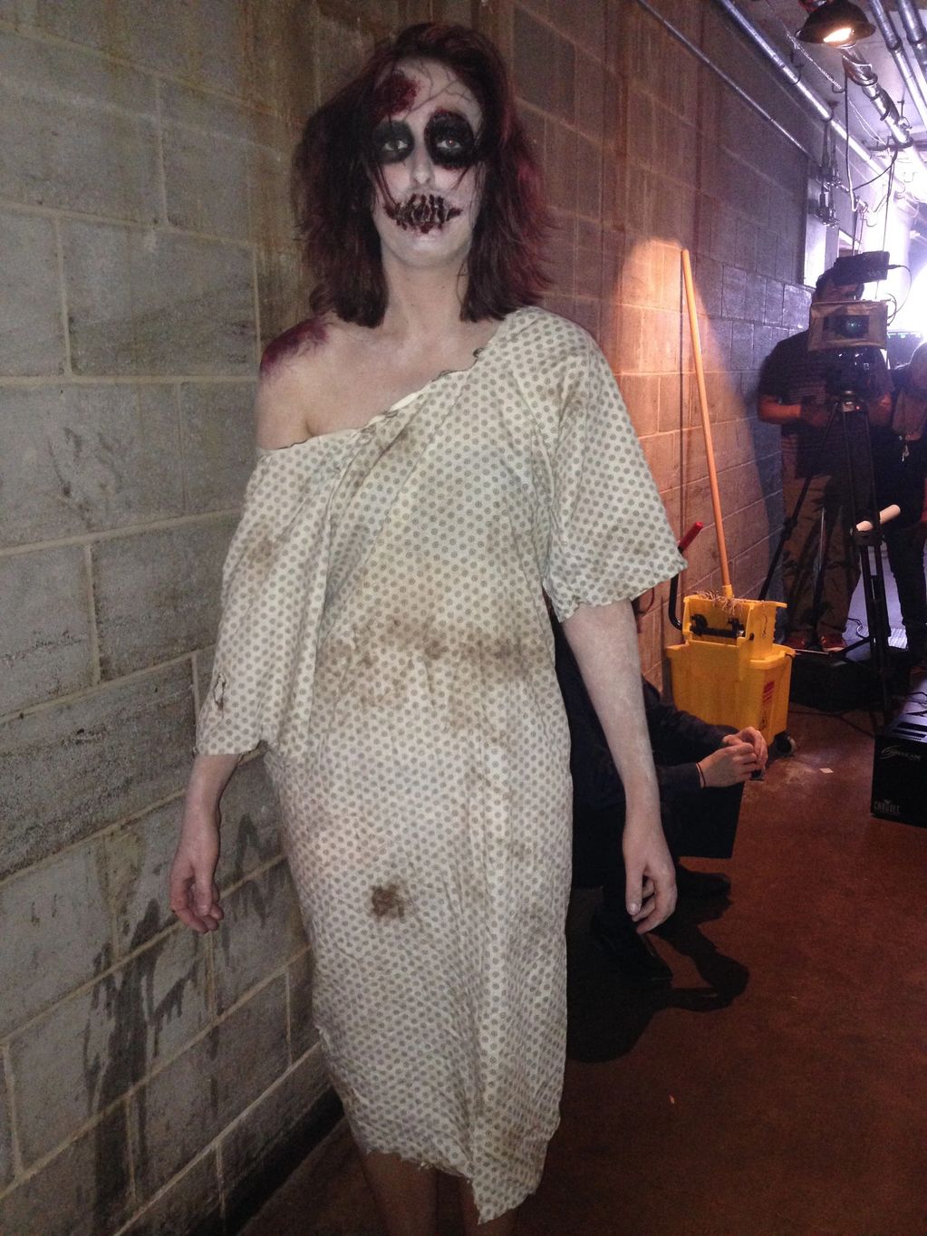 A character from a horror film wearing a dirty hospital gown standing in a dark brick hallway.
