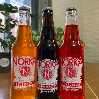 Three Norka sparkling beverage bottles in orange, root beer, and cherry-strawberry flavors.