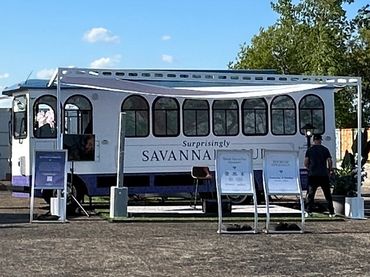 A white trolley bus labeled 'Surprisingly Savannah' with people nearby under a clear sky.
