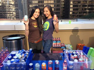 Two women smiling and holding beverages behind a table filled with assorted canned drinks.