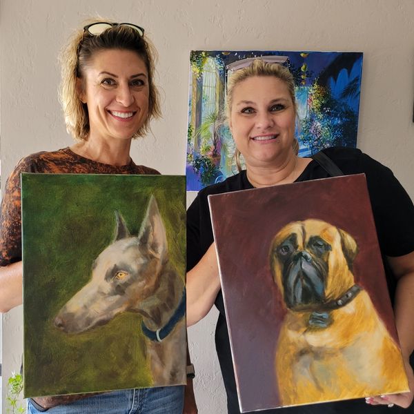 Two women proudly display their dog portraits in a cozy setting.