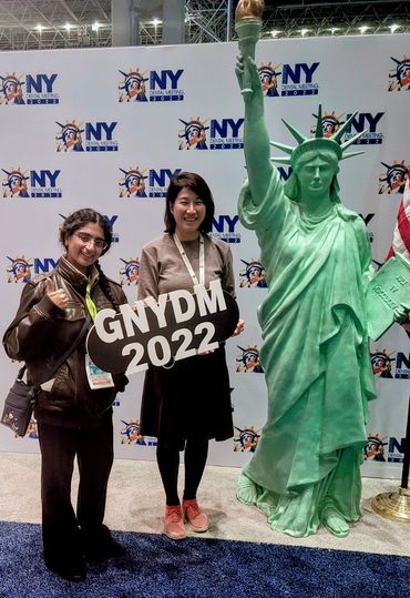 Two women smiling and holding a sign that says "GNYDM 2022" and they are standing next to a small fa