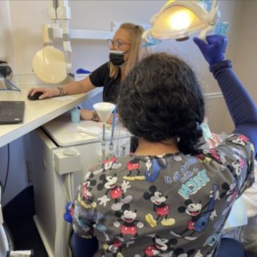 Back of a dentist who is wearing scrubs with a cartoon mouse, holding a light, and looking into a pa