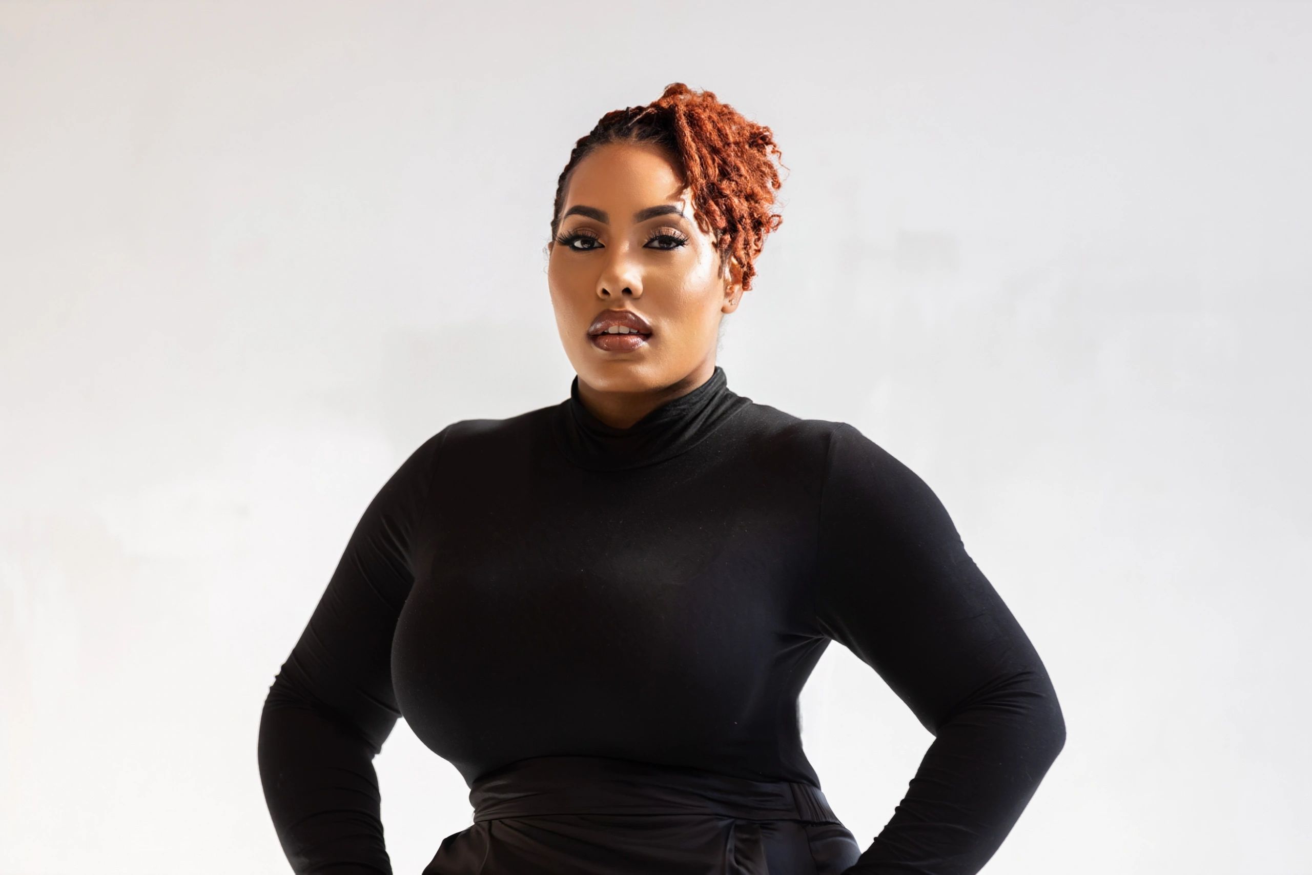 Confident woman with red curls wearing a black turtleneck posing against a plain background.