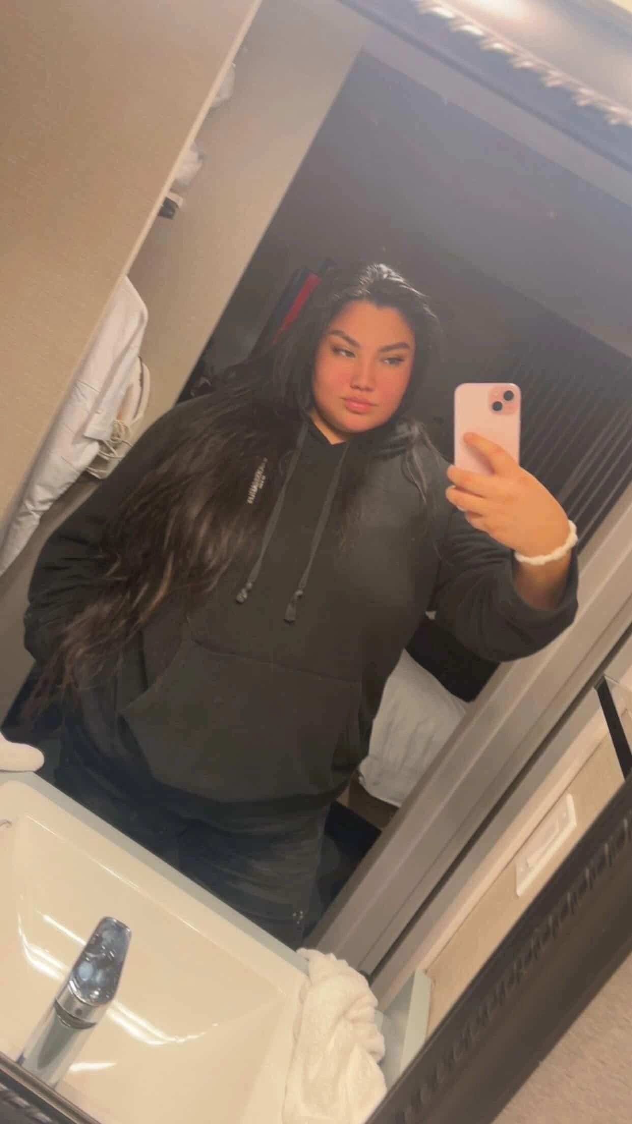 Person taking a mirror selfie wearing a black hoodie with long hair.