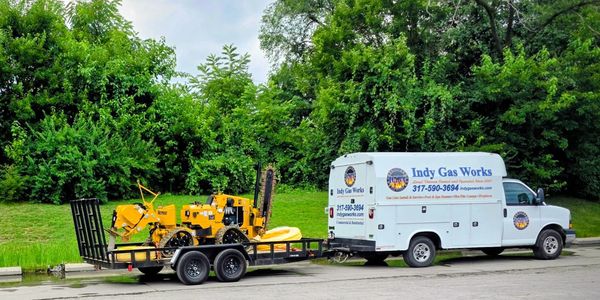 White Indy Gas Works van towing a trailer with yellow construction equipment.