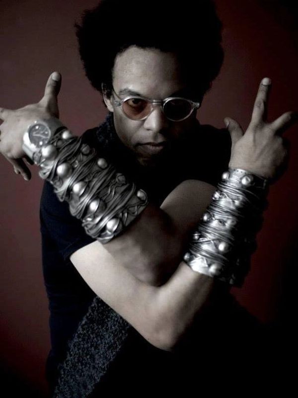 Man with afro and sunglasses wearing large, metallic arm cuffs posing with crossed arms.