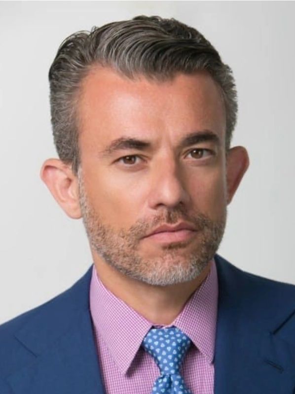 Man with gray-streaked hair, beard, wearing blue suit and patterned tie.