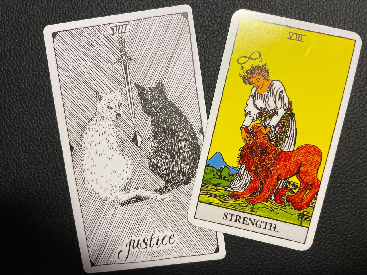 Two tarot cards. One black and white illustration of Justice (viii) with a white cat and a black cat sitting with a sword between them. Lines radiate outward in the background in a diamond pattern. The Second card is the Ryder Scott STRENGTH card with a serene woman in white, gently closing the mouth of a lion. She is crowned by a wreath of red roses, and an infinity sign hovers over her head.
