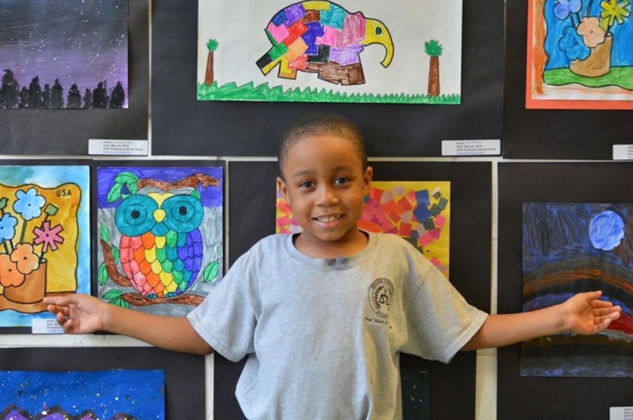 A smiling child stands in front of colorful artwork with arms outstretched.