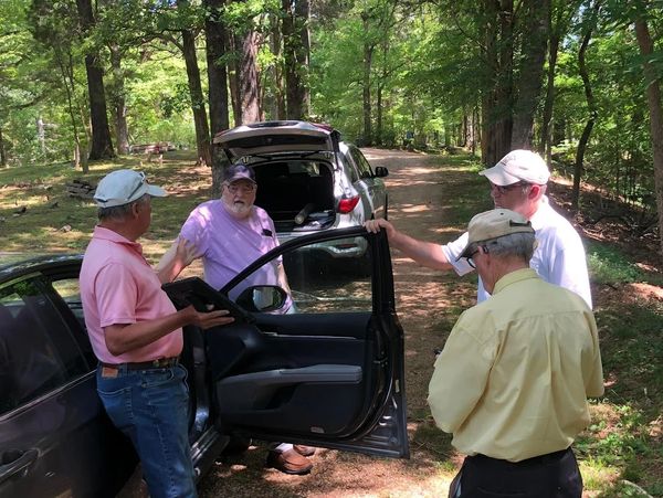 Four men in casual clothes talking near parked cars on a forest road.