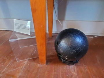 A black cannonball on the floor near a table leg and a plastic stand with a sign.