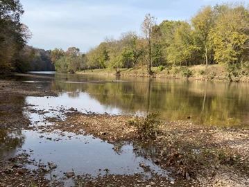 A calm river with a rocky shore and trees under a blue sky.