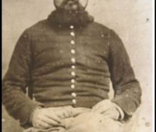 Portrait of a bearded man in vintage military uniform sitting with hands folded.