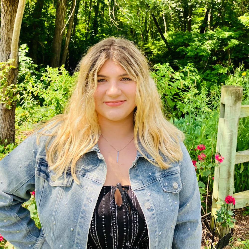 Girl with blonde hair wearing denim jacket standing outside near fence with plants behind her.