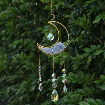 Golden crescent moon wind chime with crystal pendants hanging against green foliage.