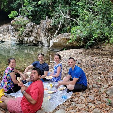 Five friends enjoying a picnic by a rocky riverbank surrounded by greenery.