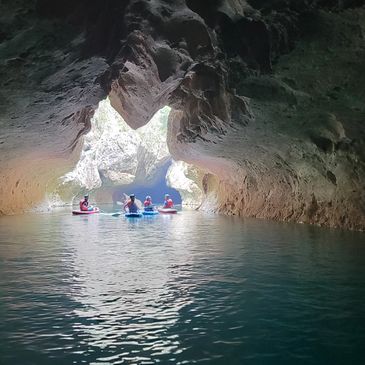 Kayakers paddle through a sunlit cave opening over calm water.