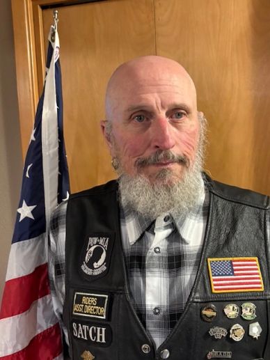 Man with a beard and leather vest with patches stands in front of an American flag.