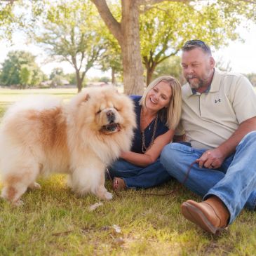 Couple sitting on grass with a fluffy dog.