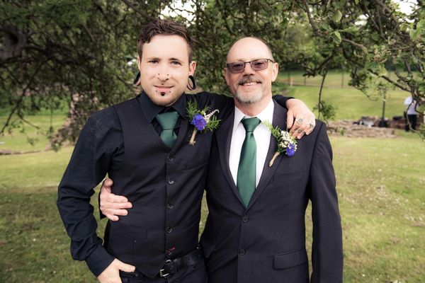 Pete with his arm round father in their wedding suits on grass and under tree