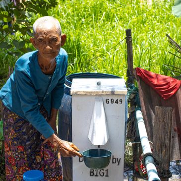 An elderly person collects water from a white water dispenser outdoors.