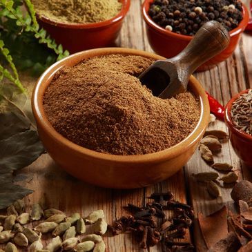 Brown spice powder in a wooden bowl with a scoop, surrounded by whole spices.