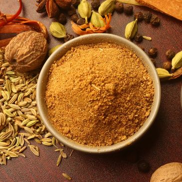 Bowl of ground spice surrounded by whole spices on a rustic surface.