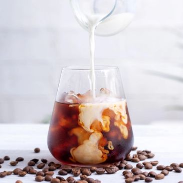 Cream being poured into a glass of iced coffee surrounded by coffee beans.