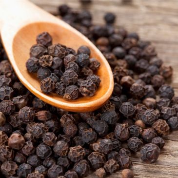 Black peppercorns in a wooden spoon and scattered on a wooden surface.