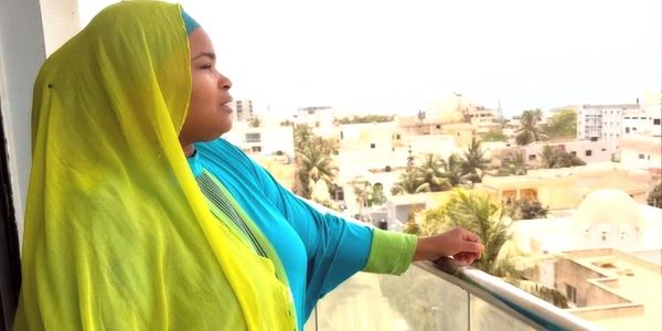 A woman in bright traditional attire gazes thoughtfully from a balcony overlooking a cityscape.