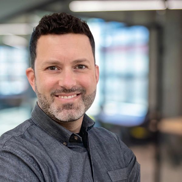 Smiling man with a short beard wearing a gray shirt in a modern office.
