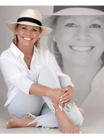 Smiling woman in a white hat and casual white outfit sitting barefoot.