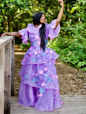 Personnage féérique en robe violette ornée de fleurs, posant gracieusement dans un décor boisé.