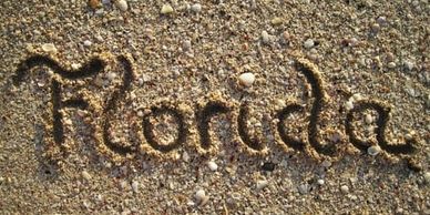 The word "Florida" written in beach sand with small sea shells scattered throughout the sand