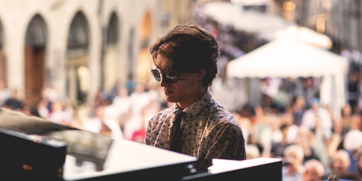 Young man in sunglasses playing piano outdoors in front of a crowd.