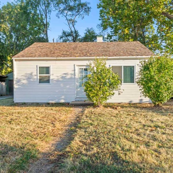 Small white house with two bushes in front and a dry lawn.