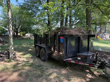 Black utility trailer filled with yard debris in a sunny backyard.